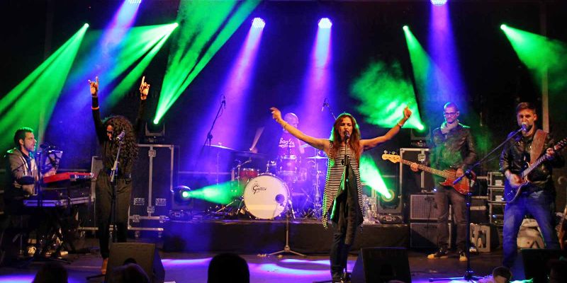 Band auf einer Showbühne mit Sängerin im vorderen Bereich. Im Hintergrund lilane und grüne Strahlen der Lichttechnik.